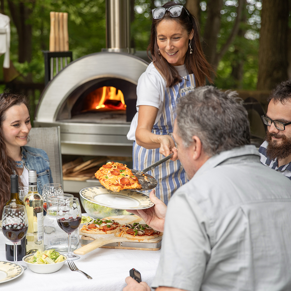 a woman takes a pizza from the oven and brings it to a convivial table of friends