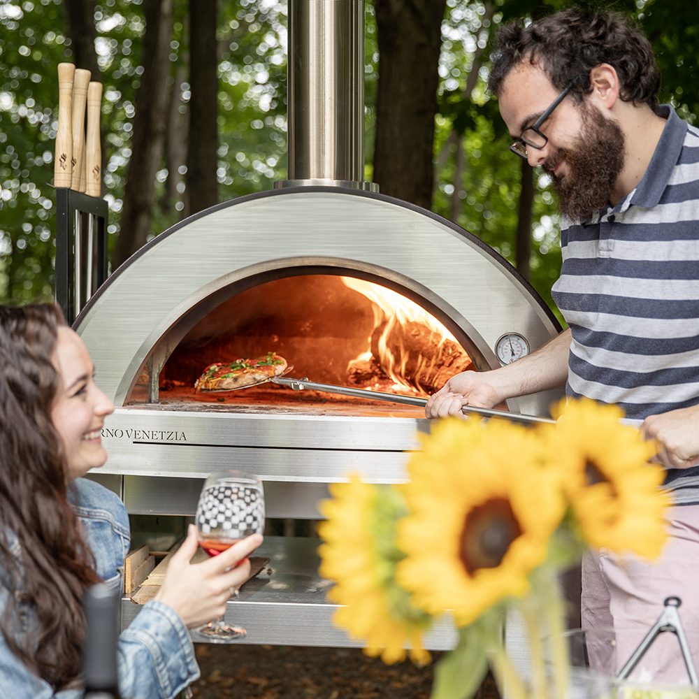 man and woman smiling in front of the oven - he bakes a pizza, she has a drink
